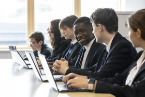 A group of Leigh Academy students are photographed sitting together at their desks, looking at their laptops and smiling.