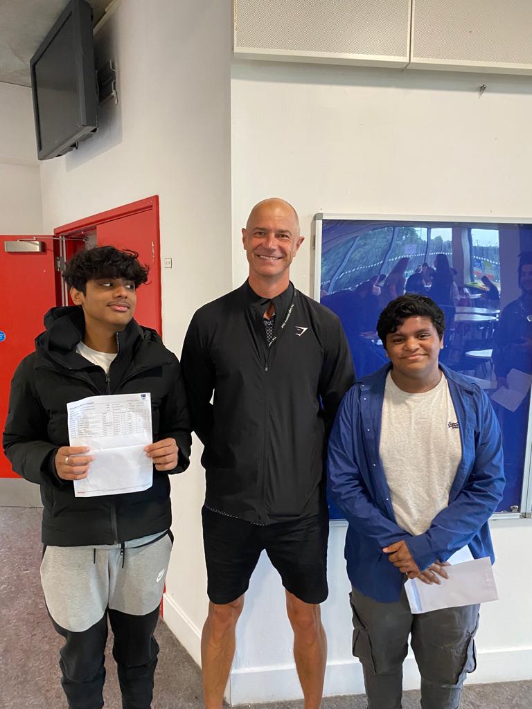 A male member of staff can be seen smiling alongside two Year 11 students who have just opened their results on GCSE Results Day 2022.