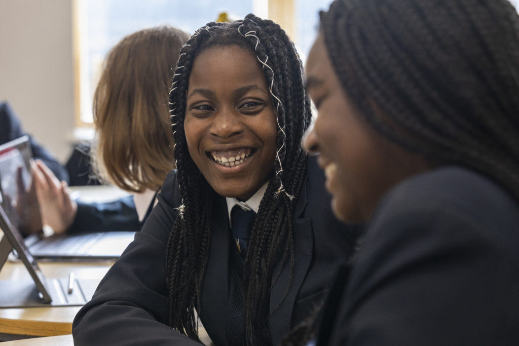 Two young girls are seen sat together at a desk, smiling at one another.