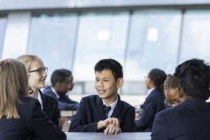 A young male Year 7 student is seen sat at a desk, smiling and surrounded by his peers in a dining area.