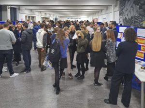 A large group of Post-16 students are seen gathered in an atrium area of the academy building for an open evening presentation.