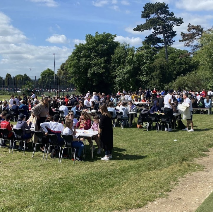 A large crowd of both students and staff are seen sat on chairs and tables outdoors on the academy grounds. A bright blue sky is seen above them.