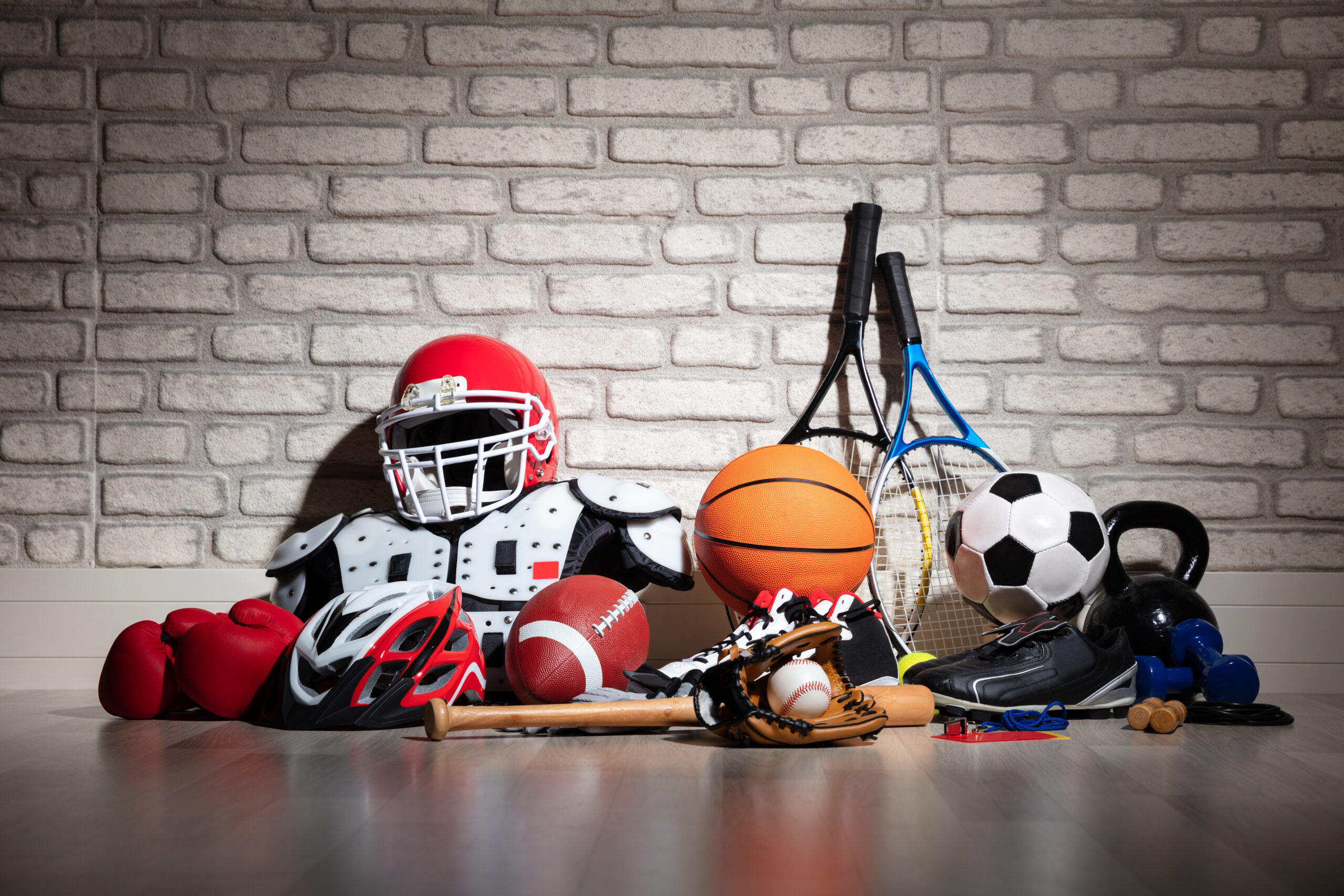 Assorted sporting equipment from various different games is seen stacked in a pile on the floor, against a grey brick wall.