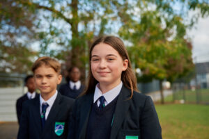A girl and boy are seen in the foreground of the photo, smiling for the camera whilst wearing their Leigh Academy uniform.