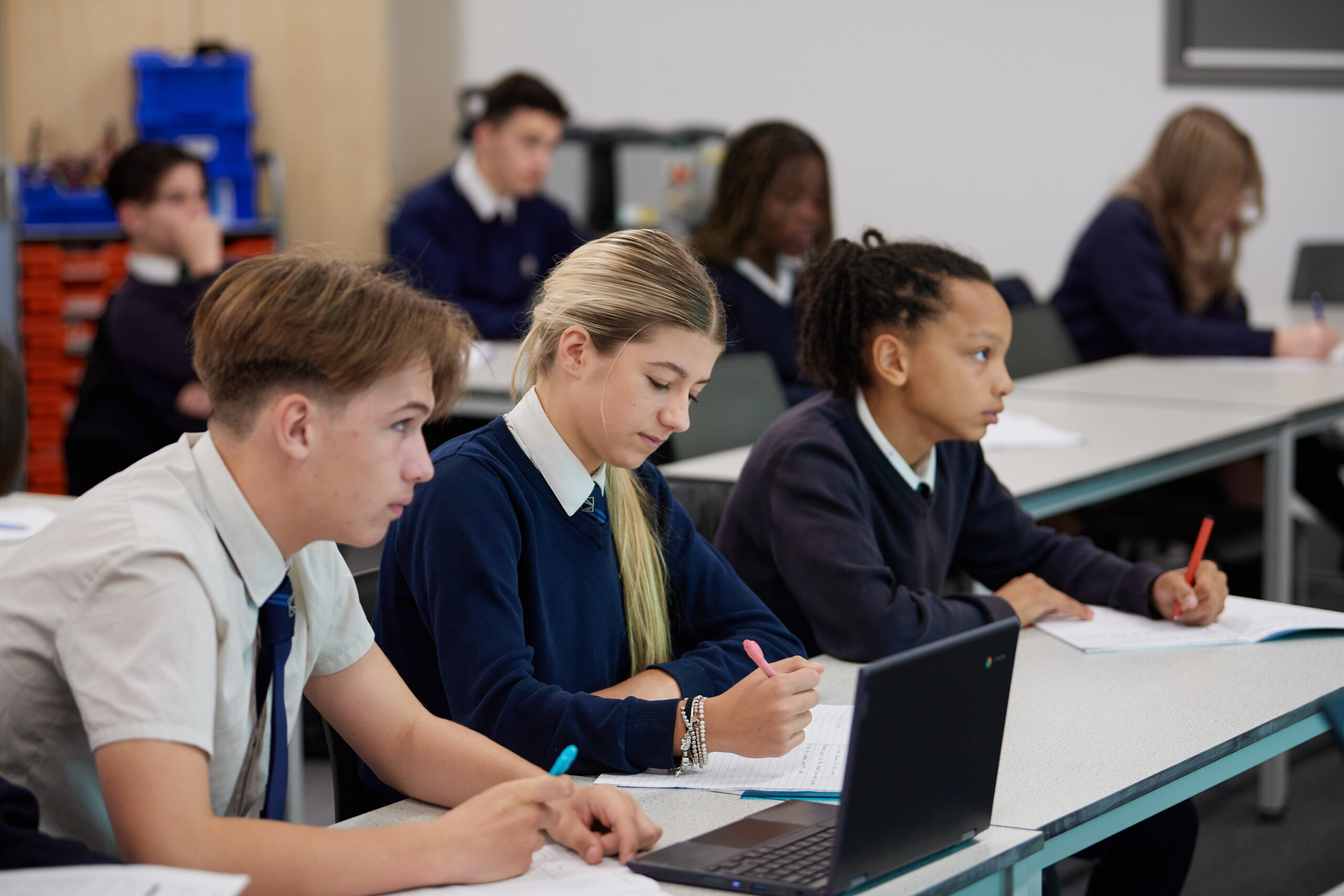 Three Leigh Academy students, one boy and two girls, are pictured working at their desks and looking towards the front of the classroom.