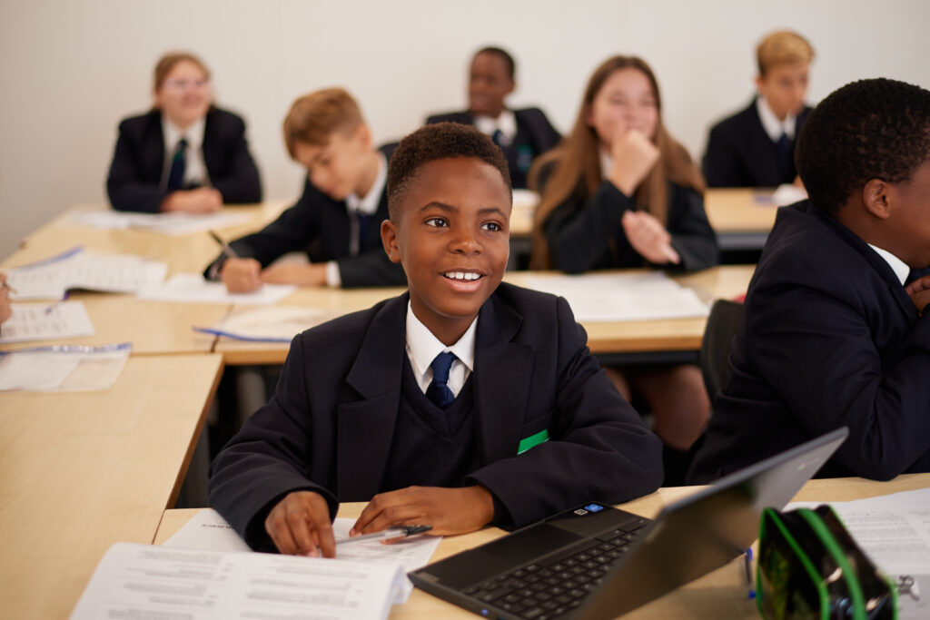 A young boy is seen wearing his academy uniform and smiling for the camera during a lesson, surrounded by his peers.