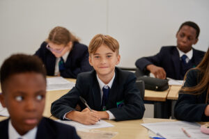 A young, male Leigh Academy student is pictured in the foreground, sat at his desk with a pen in his hand and smiling at the camera.